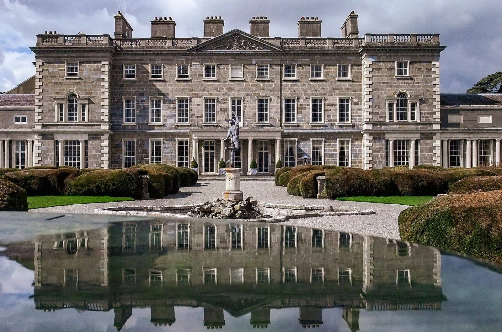 The grand stone front of Carton House mirrored in a still pool, with clipped hedges and a statue on a fountain in front.