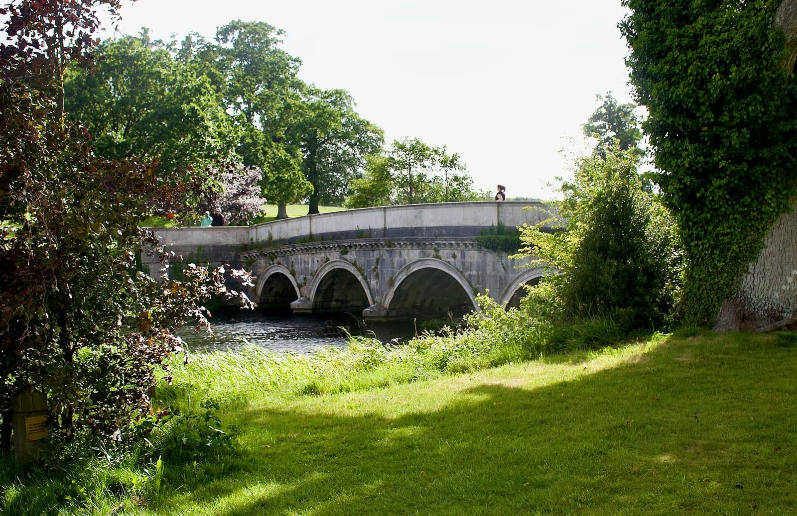 A stone bridge of several arches crosses the Rye Water in the Carton demesne, framed by tall trees and a sunlit lawn.