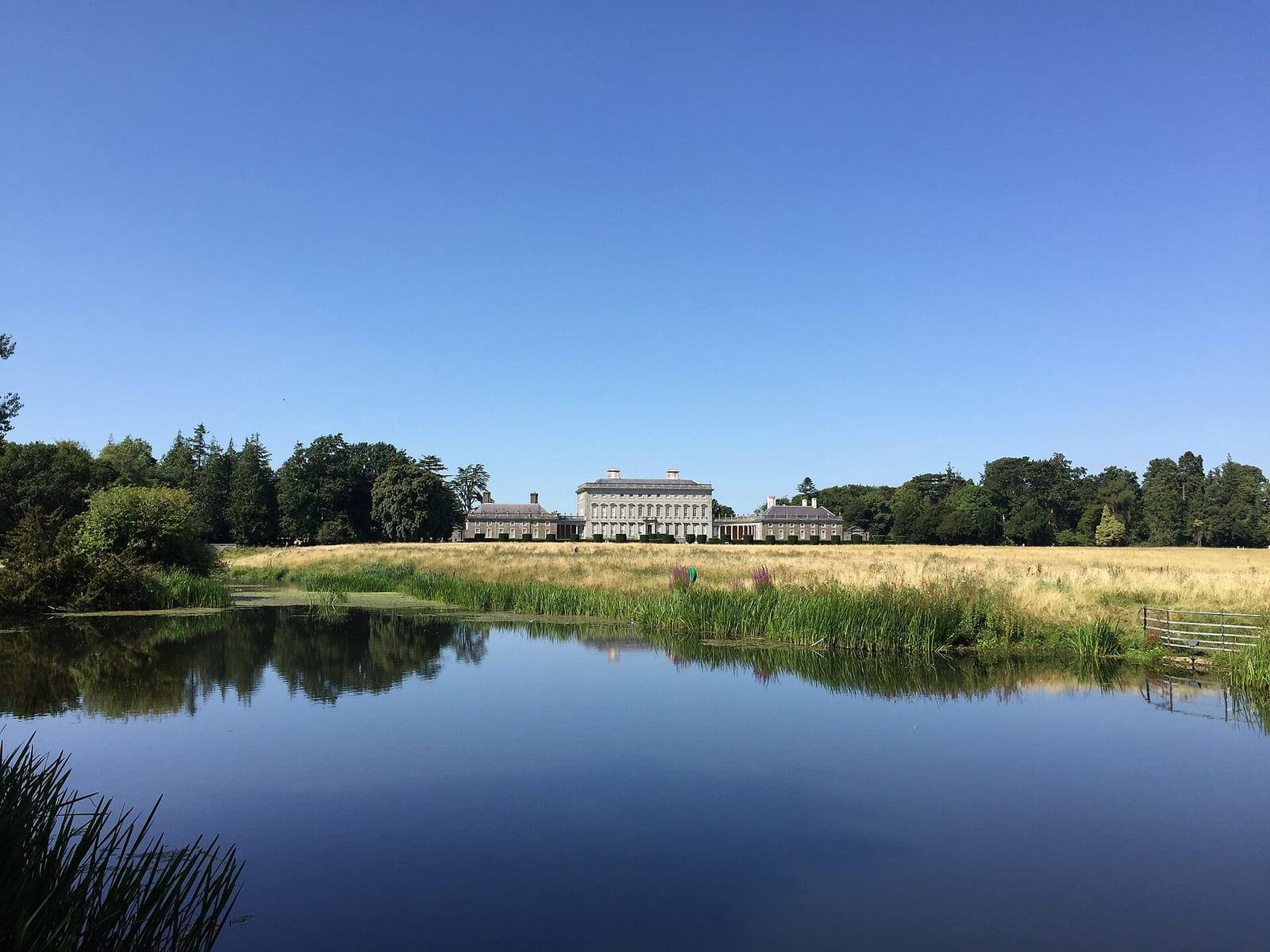 Castletown House, a long Palladian mansion, seen across a still, deep-blue lake and a meadow under a clear blue sky.