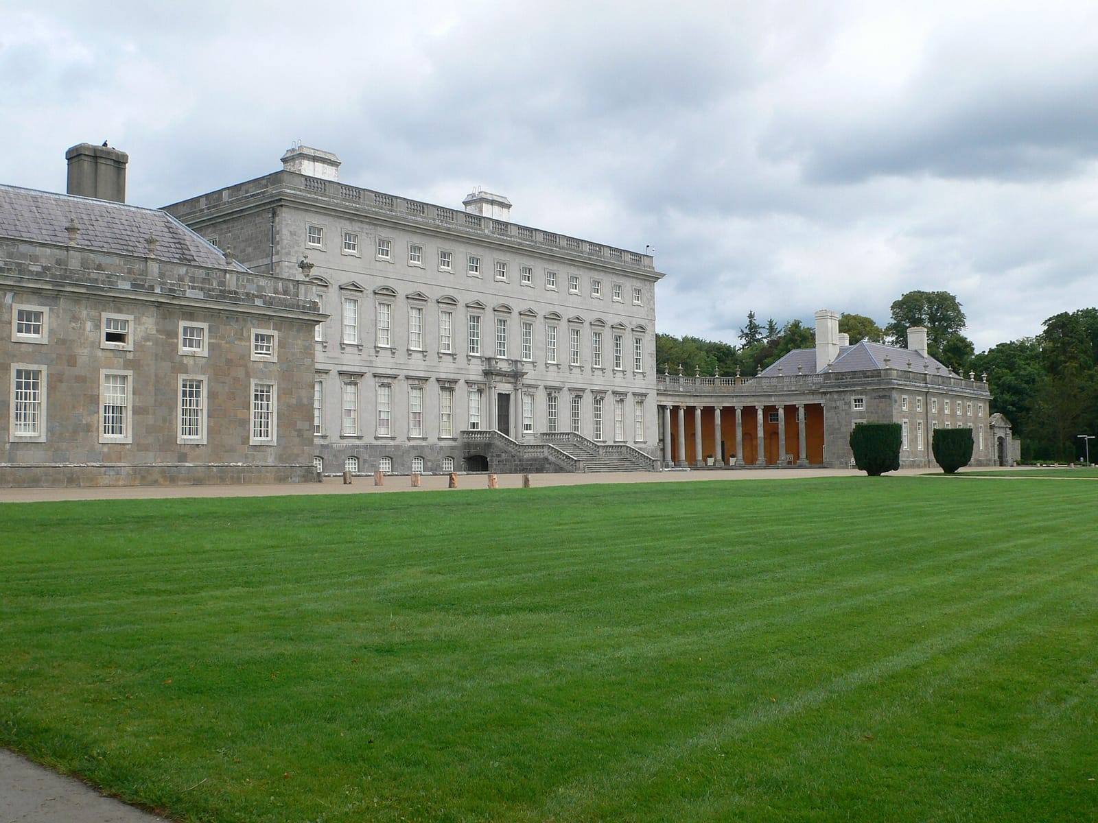 The grey stone front of Castletown House and its curved colonnade, behind a wide, freshly mown lawn on a cloudy summer day.