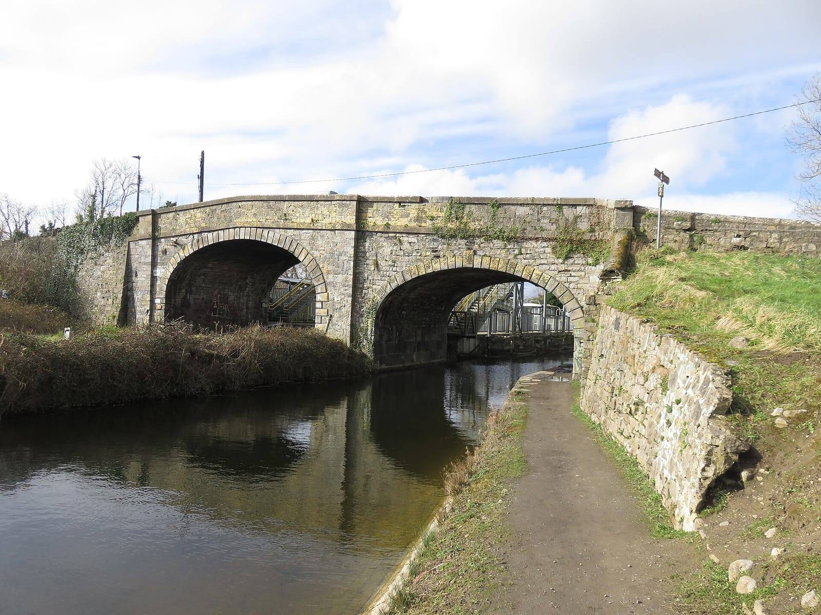The double stone arch of Cope Bridge over the Royal Canal at Confey, with a grassy towpath and a rough stone wall on the right.
