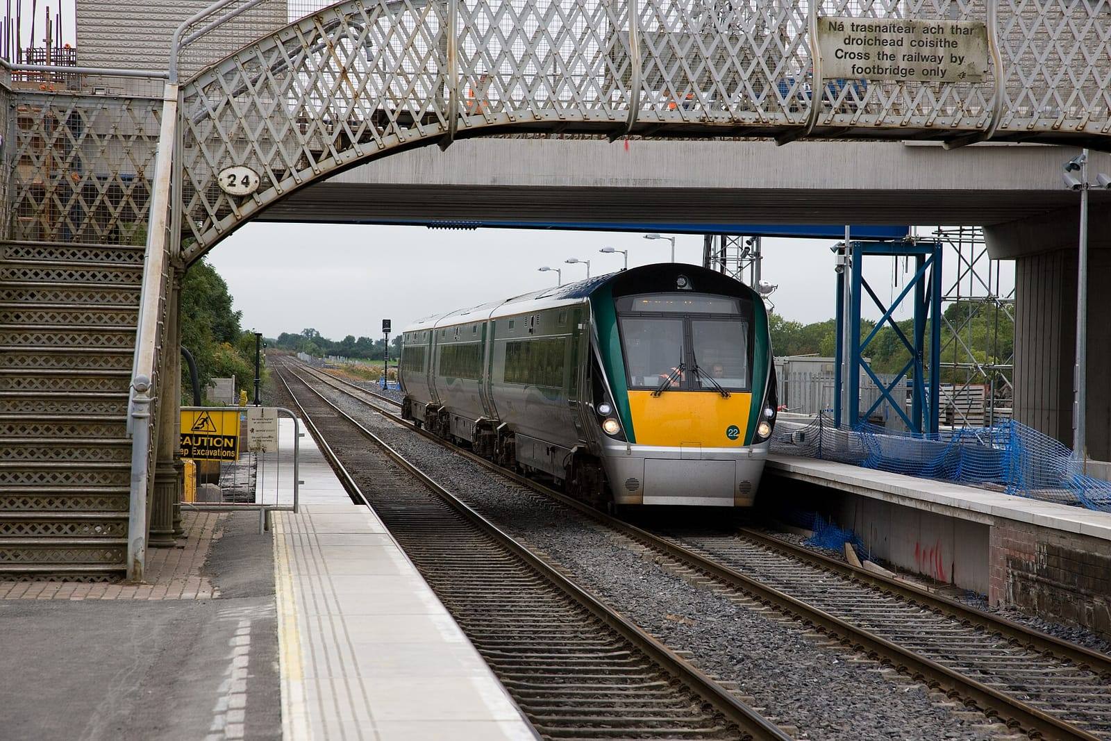 A green and yellow Irish Rail Intercity train arrives at Hazelhatch and Celbridge station beneath an ornate iron footbridge.