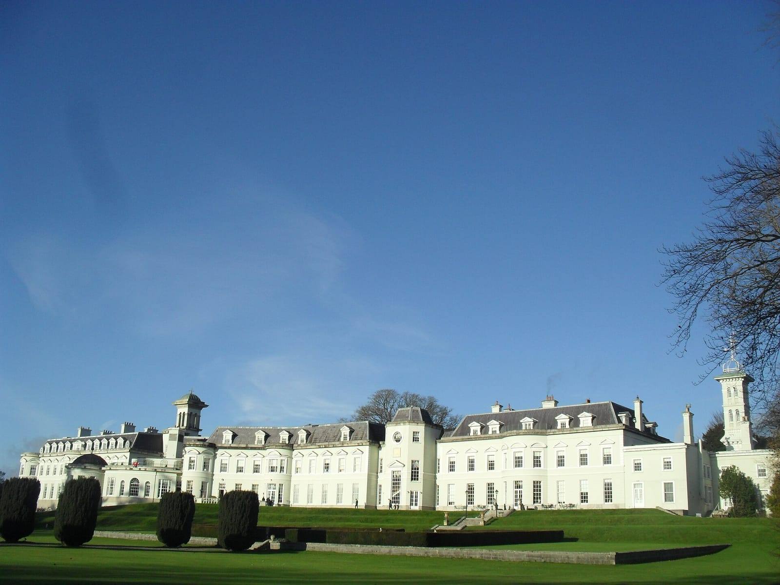 The long white front of the K Club hotel at Straffan behind clipped yew trees and a lawn, under a deep blue sky.