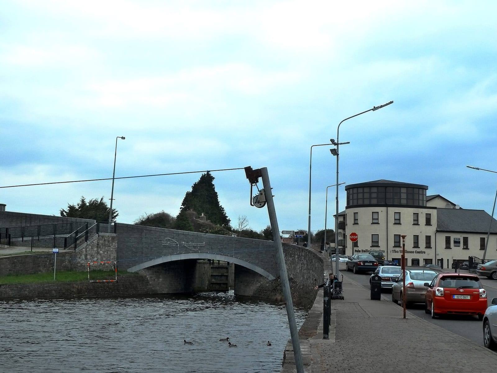 The Royal Canal in Kilcock, with ducks on the water below a stone road bridge and cars parked along the street on the right.