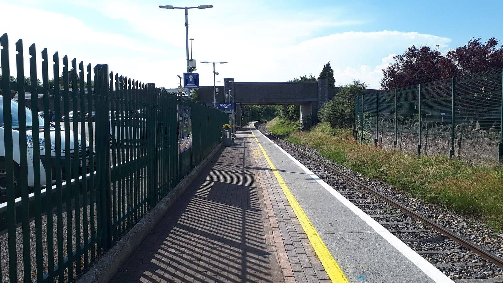 The platform at Kilcock station, with a yellow safety line, a tall green fence and a road bridge in the distance under a blue sky.