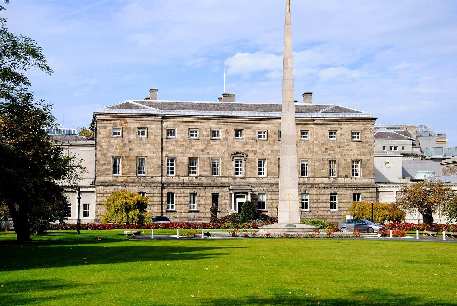 The stone front of Leinster House in Dublin seen across a green lawn, with a tall obelisk in front and flower beds below.