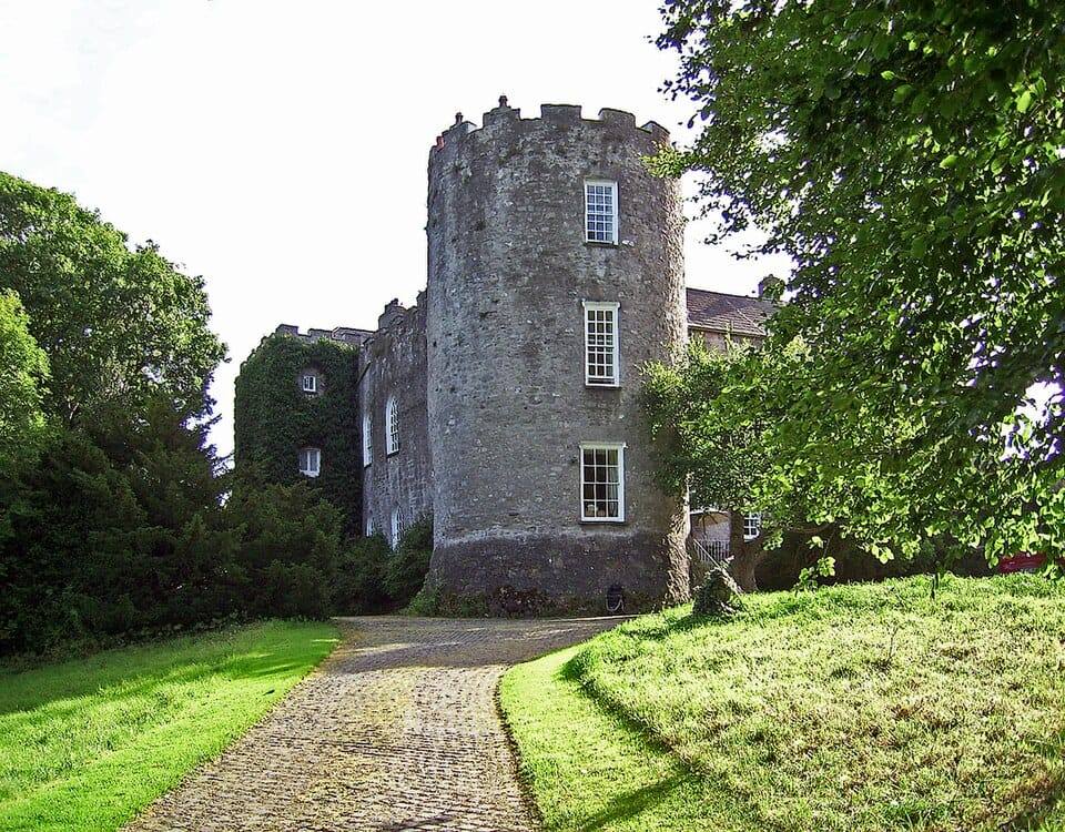 The grey stone round tower of Leixlip Castle and its ivy-covered wing, at the top of a cobbled avenue between grassy banks and trees.