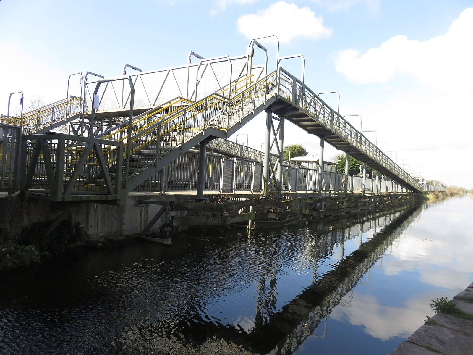 The long metal footbridge of Leixlip Confey station reflected in the still water of the Royal Canal under a blue sky with white clouds.