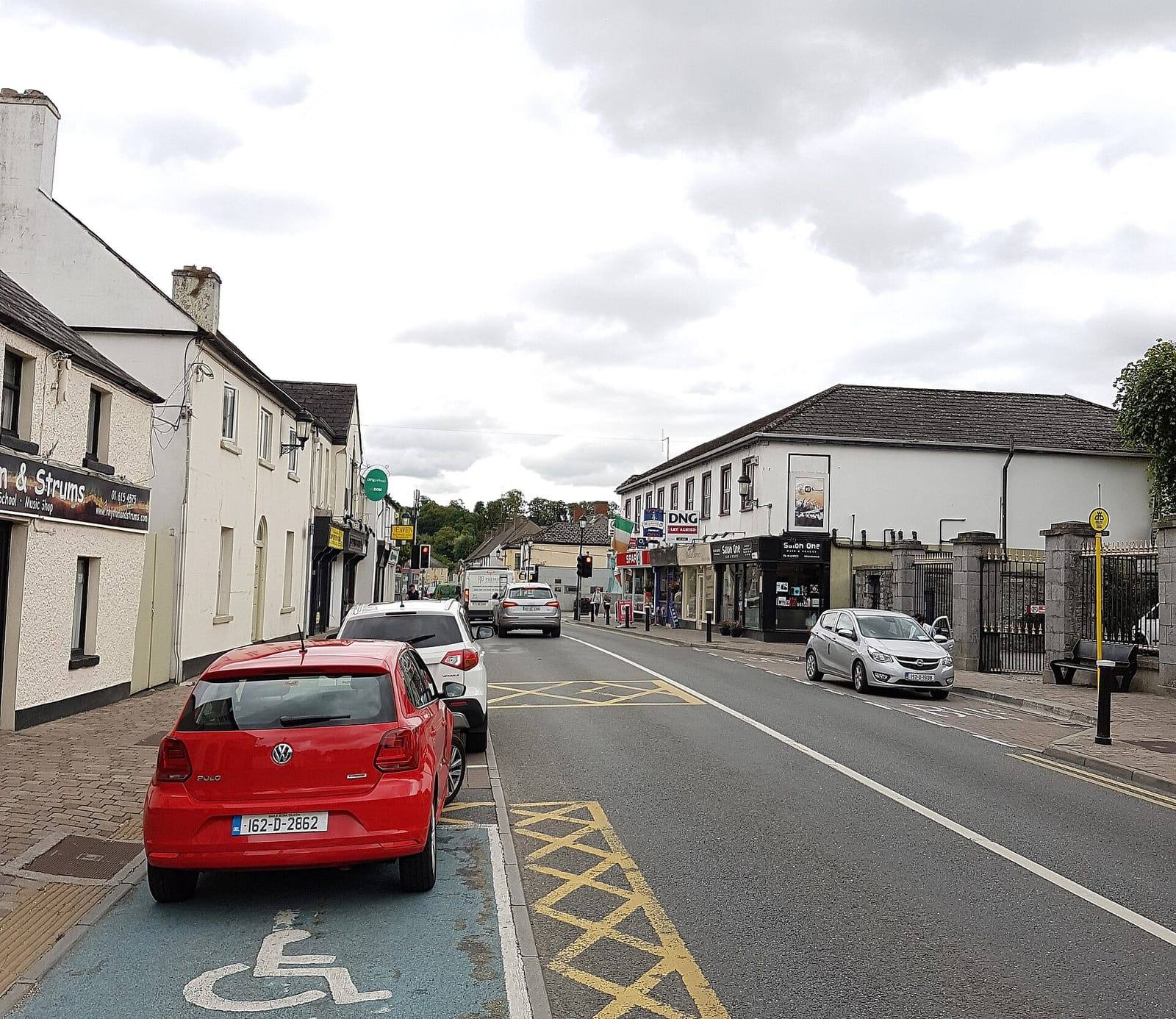 Leixlip's Main Street under a cloudy sky, with white two-storey shopfronts on both sides and cars parked along the kerb.