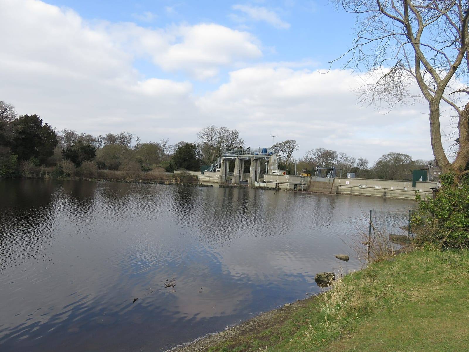 The broad, still reservoir on the Liffey at Leixlip, with the hydroelectric dam's sluice building on the far bank under a bright, cloudy sky.