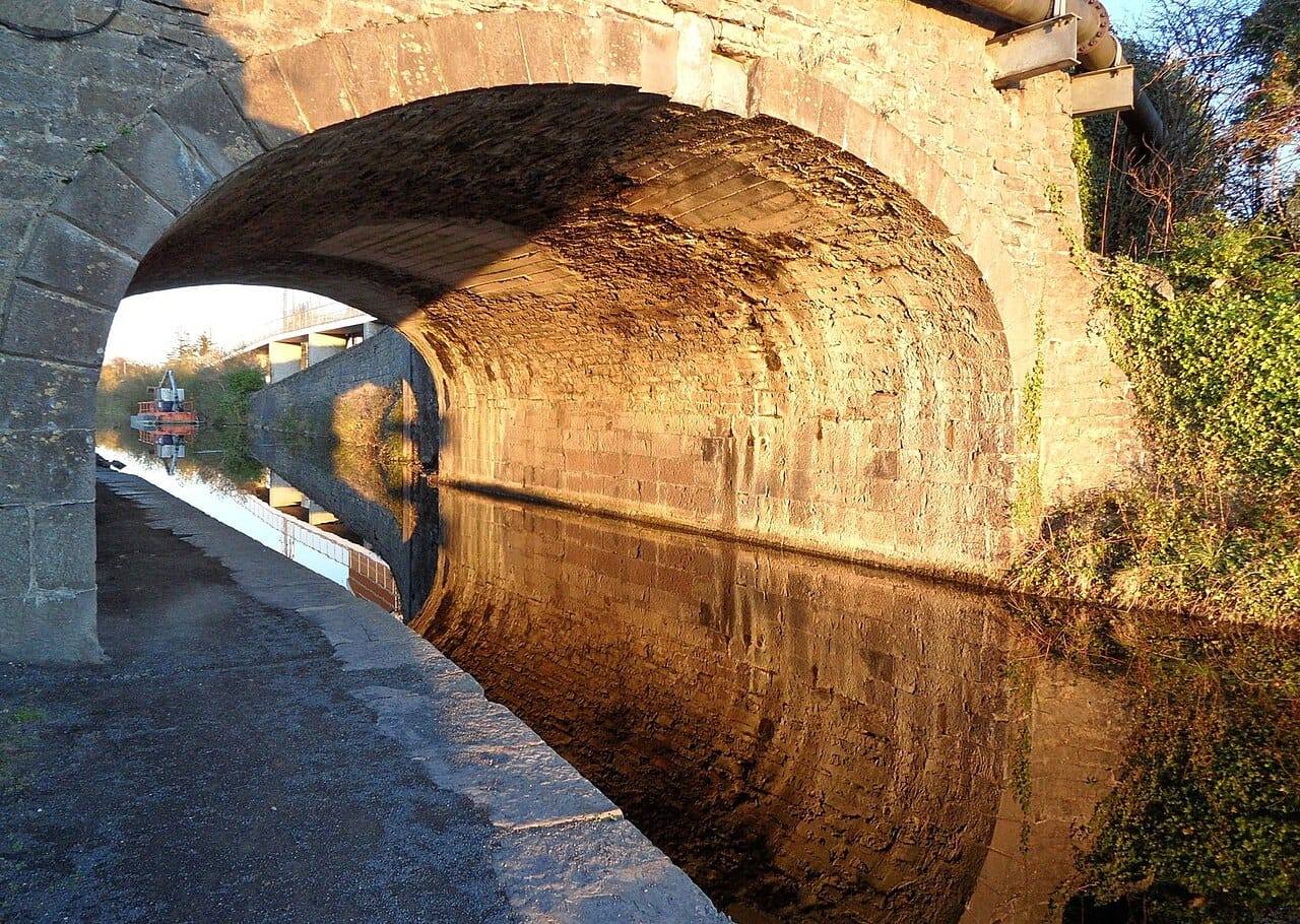 Low sun lights the stone arch of Louisa Bridge, its underside glowing orange and reflected in the Royal Canal beside the towpath.