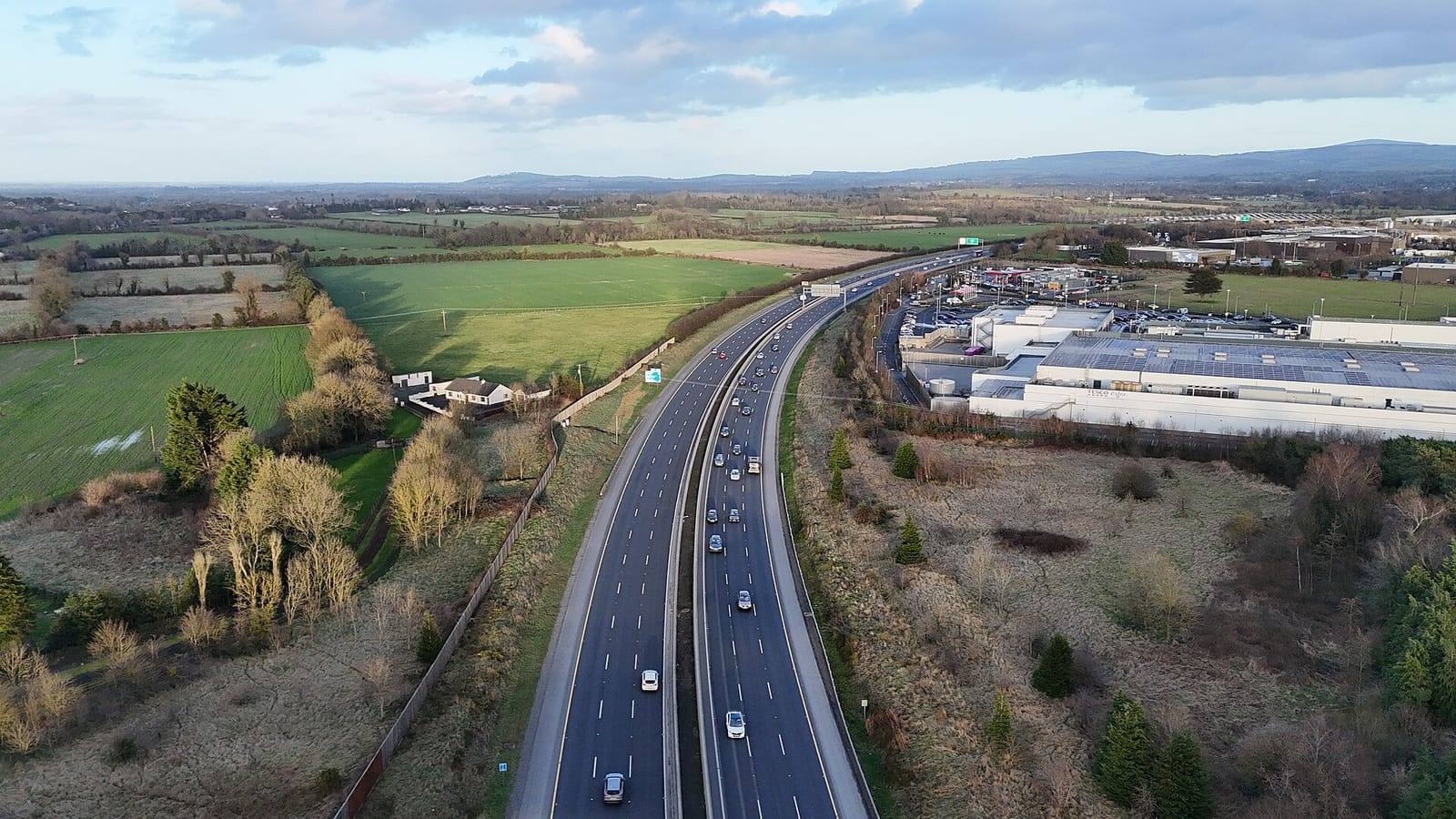 An aerial view of the M7 motorway curving past Naas, with traffic on both carriageways, an industrial estate on the right and fields stretching to the hills.