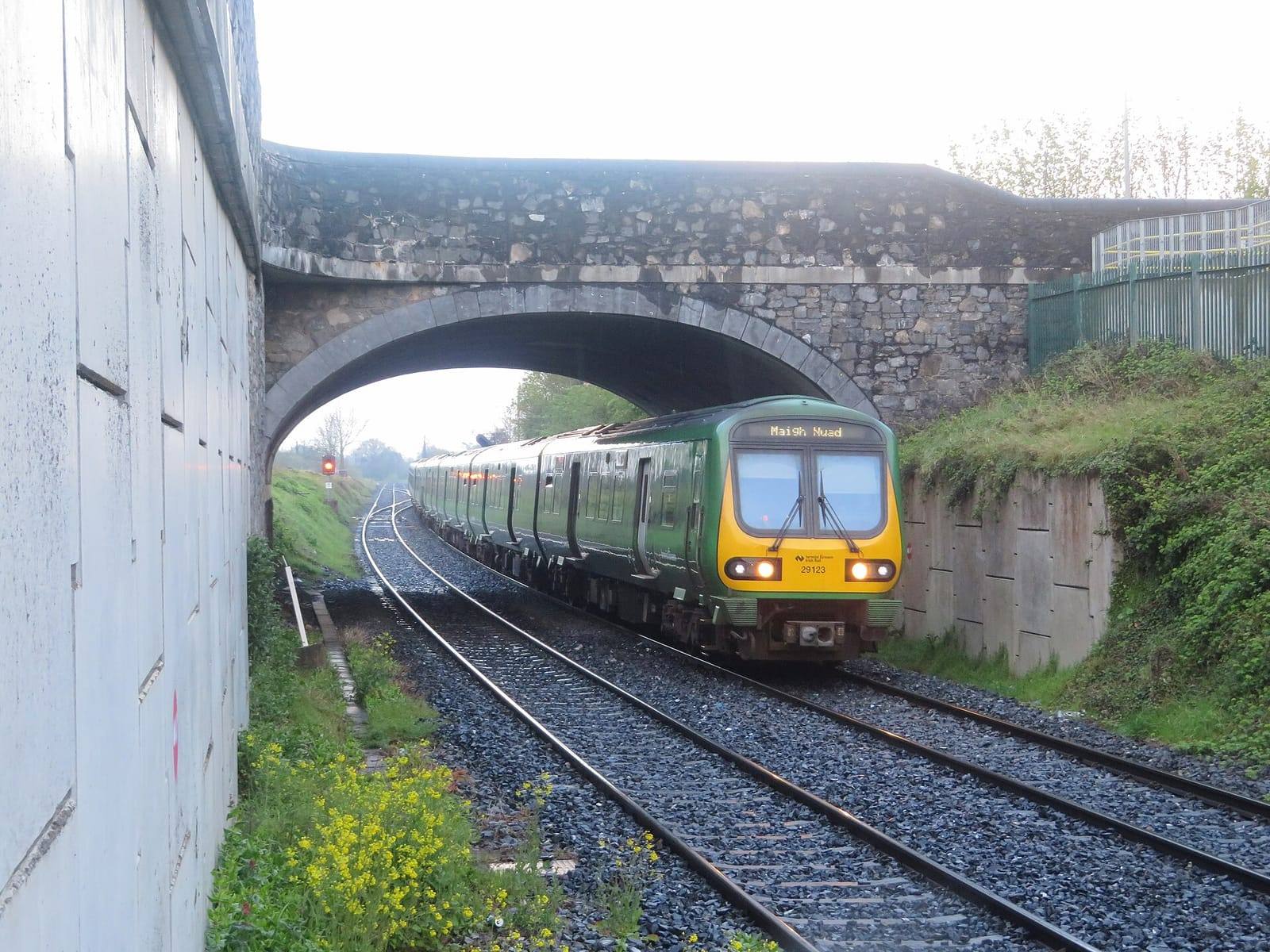 A green and yellow Irish Rail commuter train passes under a stone road bridge on its way into Maynooth, beside a tall concrete wall.