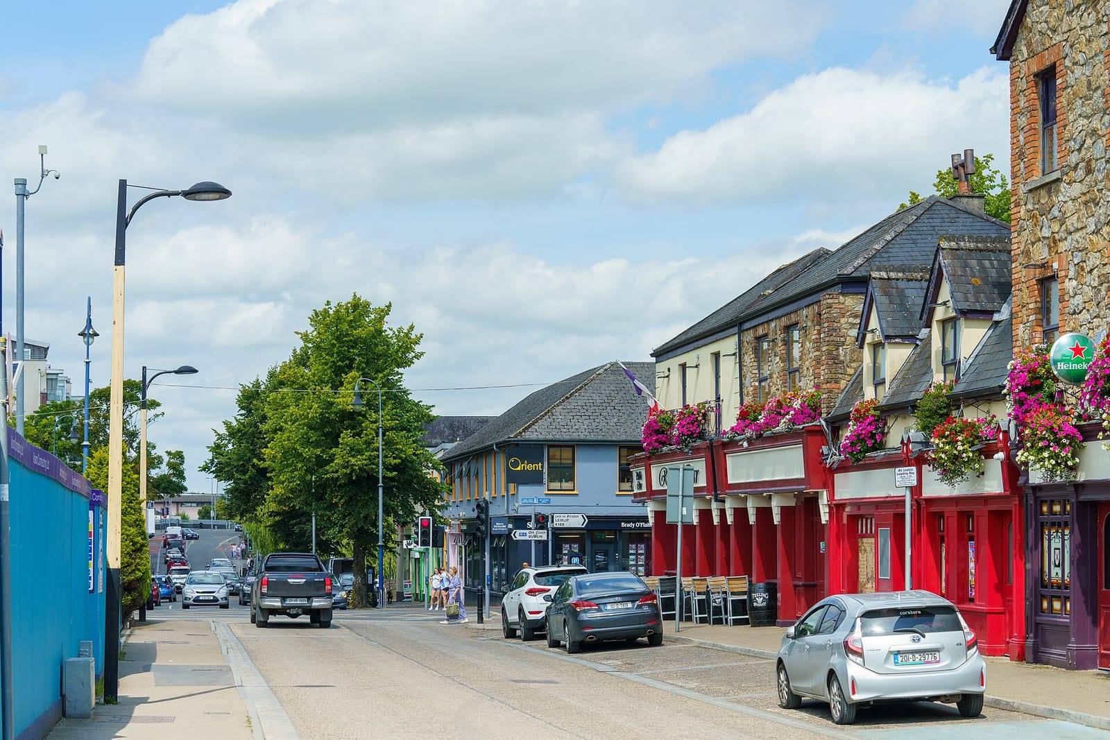 Leinster Street in Maynooth on a summer day, with hanging baskets on a red-fronted pub, shops along the street and traffic heading into town.