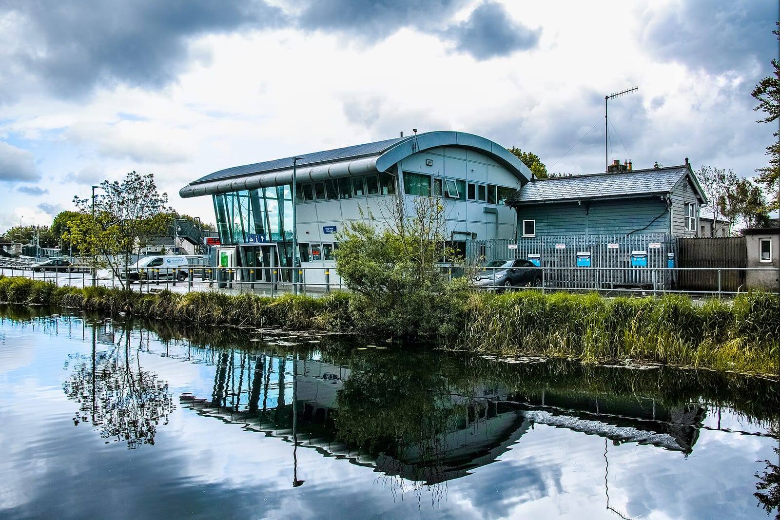 Maynooth's curved glass and steel station building reflected in the still water of the Royal Canal harbour under a dramatic, cloudy sky.