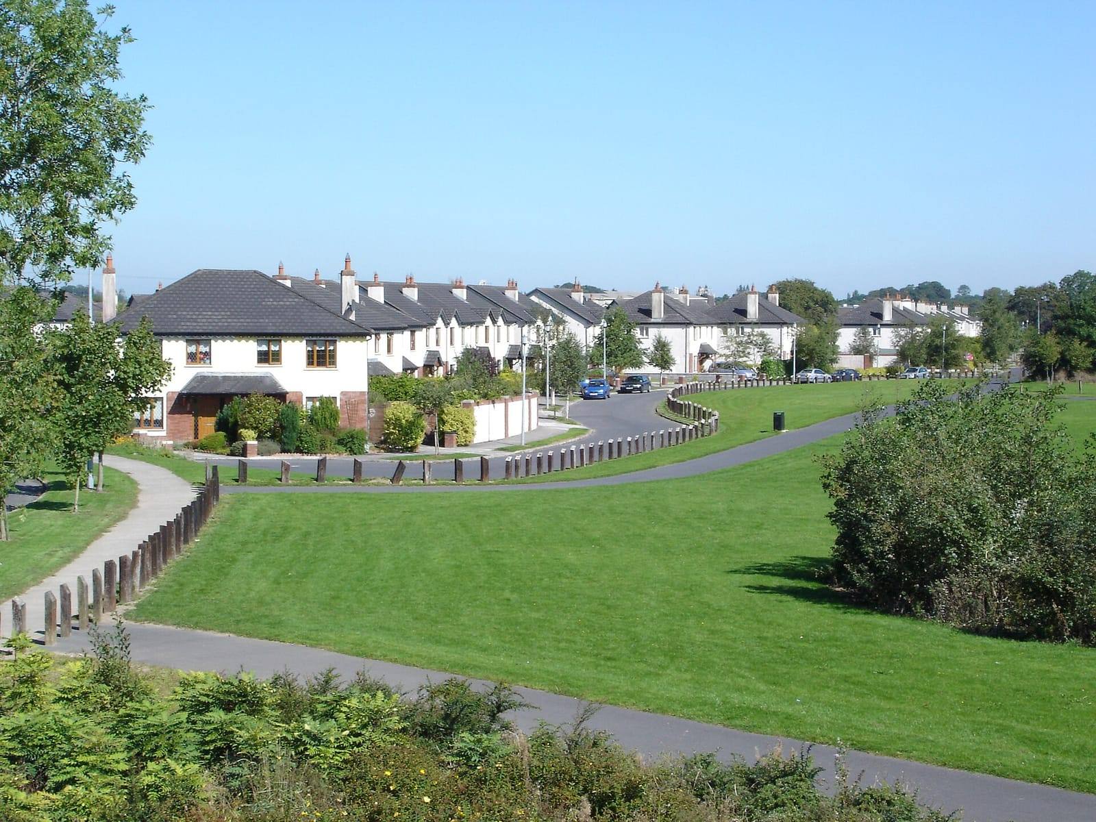 Rows of white two-storey houses curving around a large green with a path through it, under a clear blue sky.