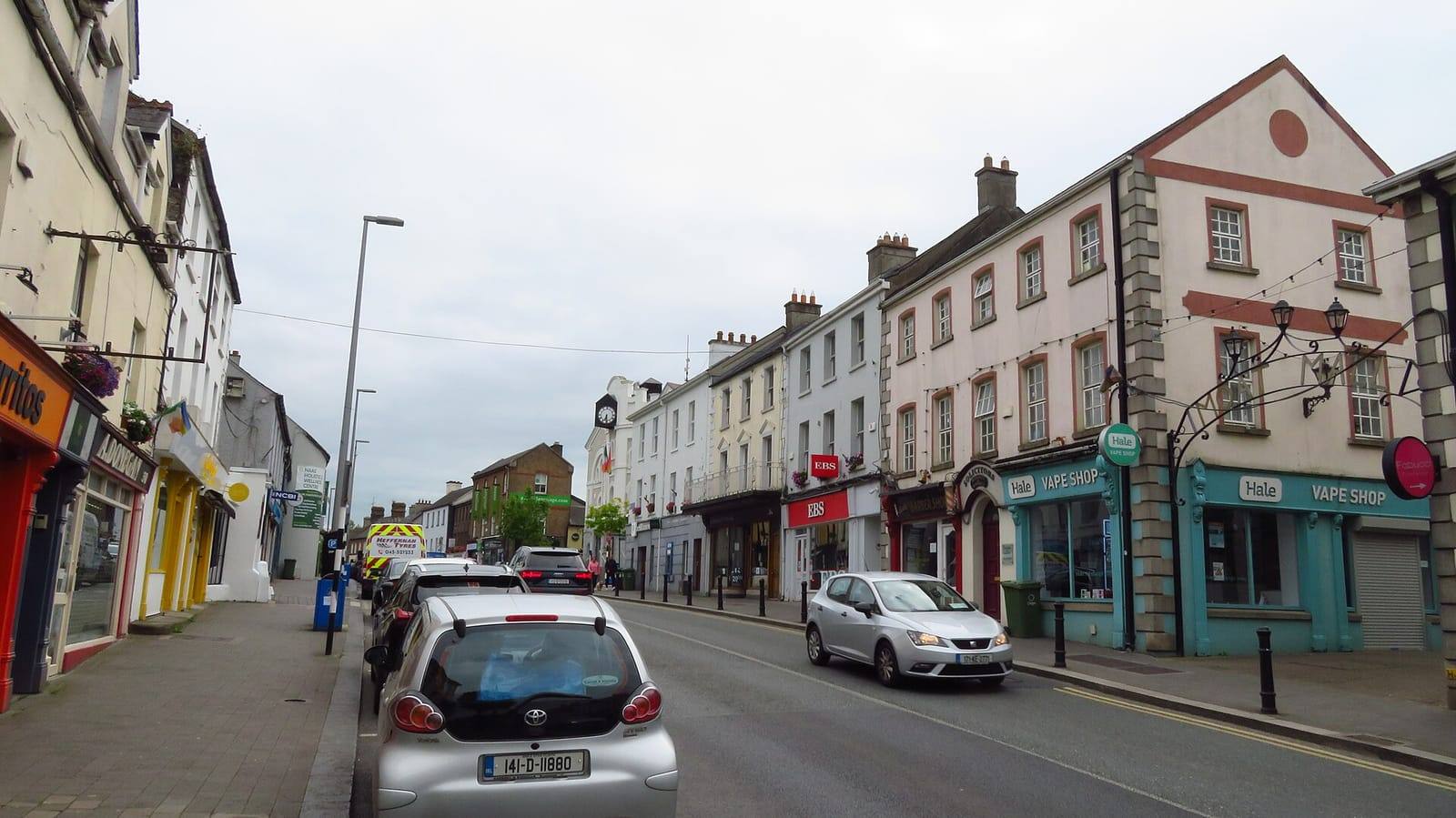 North Main Street in Naas, with painted three- and four-storey buildings and shopfronts on the right and parked cars along the street.