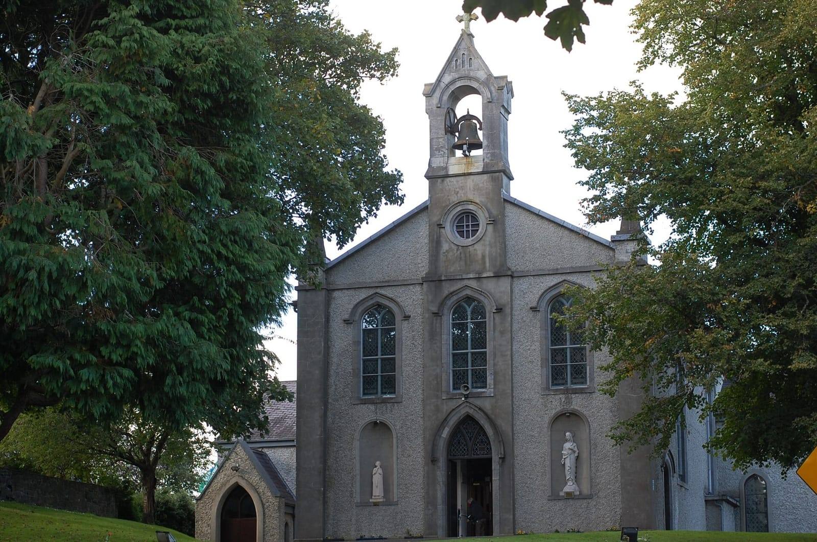 The pale grey front of the Church of Our Lady's Nativity in Leixlip, with a small bell tower and statues in niches, between tall trees.