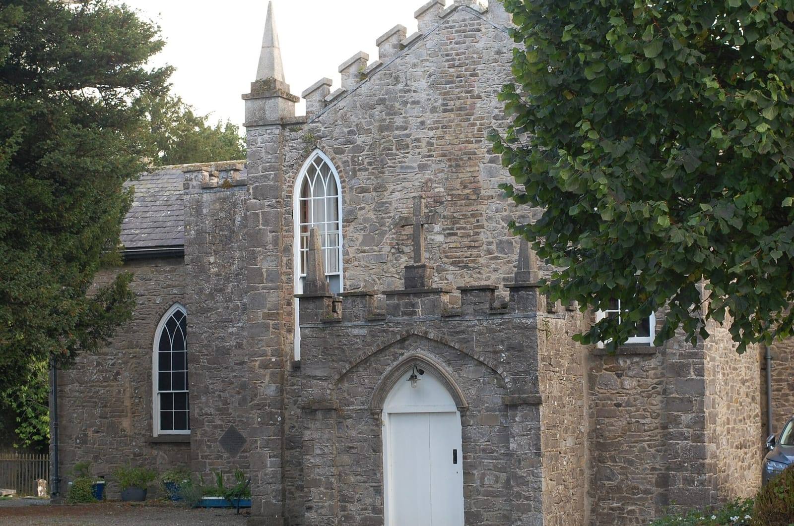 The grey stone gable of St Mary's Church in Leixlip, with a battlemented porch, a white arched door and tall pointed windows, framed by trees.
