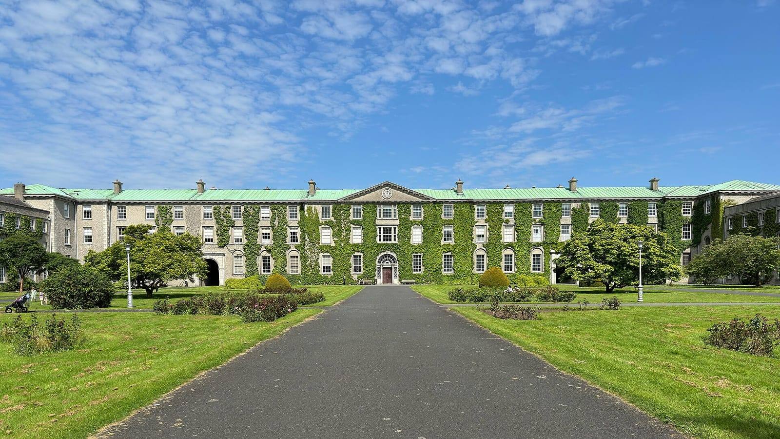 A long, straight drive between lawns leads to the ivy-covered front of St Patrick's College, Maynooth, under a blue sky with light cloud.