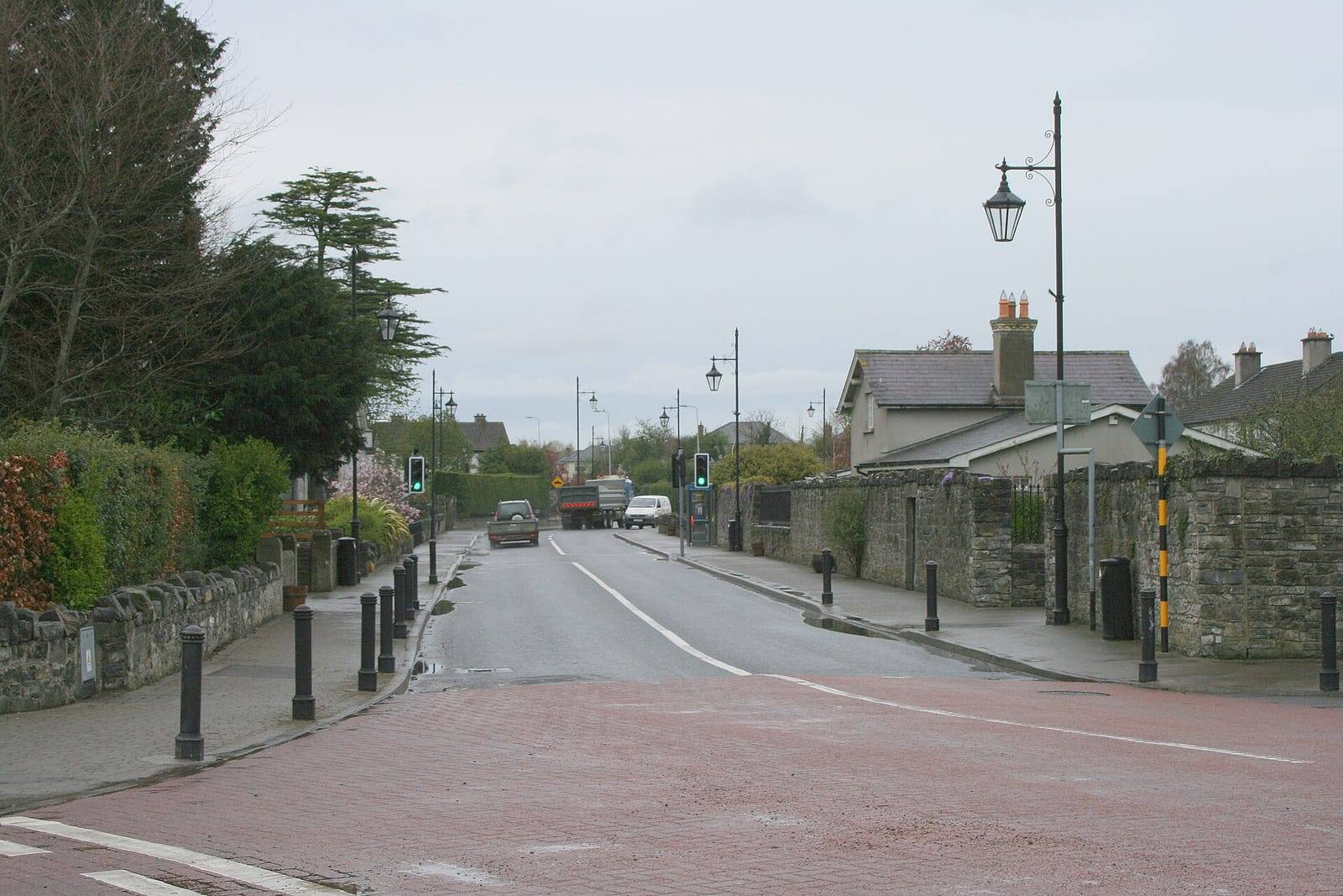 A quiet road through Straffan village, lined with stone walls, street lamps and bollards, on an overcast day.